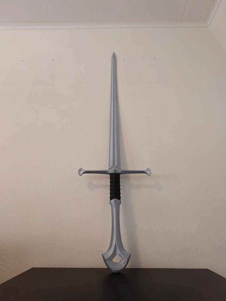 May include: A silver-colored sword with a long, pointed blade and a crossguard. The handle is black and gray, and the sword is displayed against a white wall. The sword's overall design suggests a fantasy or historical theme.