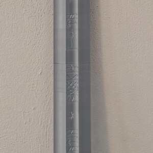 May include: A vertical, gray, textured object with embossed text. The text appears to be a series of words and symbols. The object is set against a neutral, off-white wall.