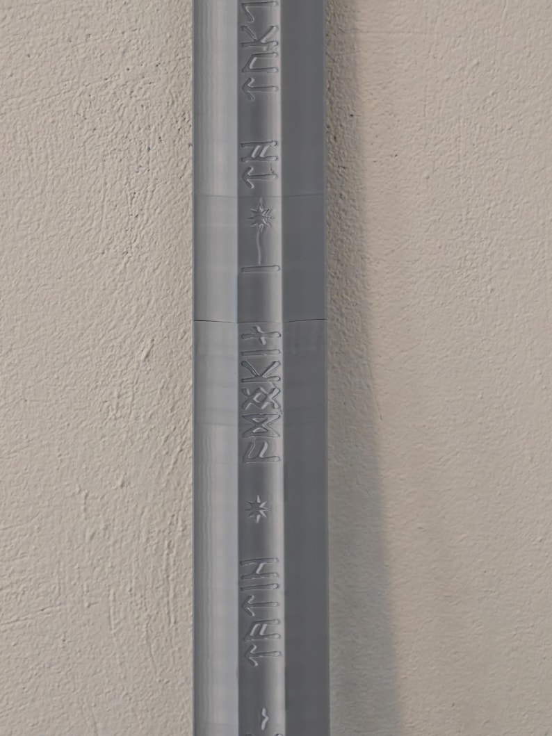 May include: A vertical, gray, textured object with embossed text. The text appears to be a series of words and symbols. The object is set against a neutral, off-white wall.