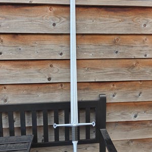 May include: A silver-colored sword with a long, pointed blade and a cross-shaped hilt. The sword is displayed upright against a wooden background and a black bench. The hilt has a dark gray grip and decorative elements.