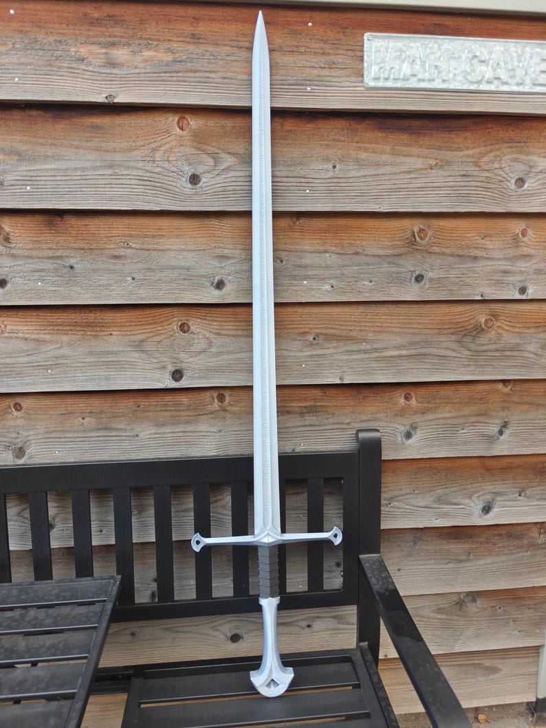 May include: A silver-colored sword with a long, pointed blade and a cross-shaped hilt. The sword is displayed upright against a wooden background and a black bench. The hilt has a dark gray grip and decorative elements.