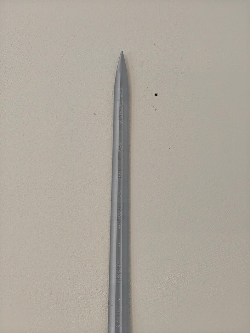 May include: A silver-colored knitting needle with a pointed tip. The needle has measurements etched along its length. The background is a neutral, off-white color.
