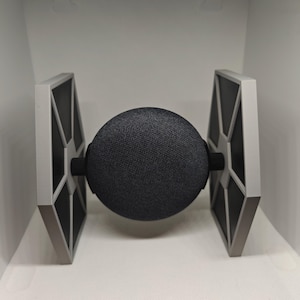 Puede incluir: Un altavoz inteligente Google Home negro con un soporte TIE fighter blanco y gris.