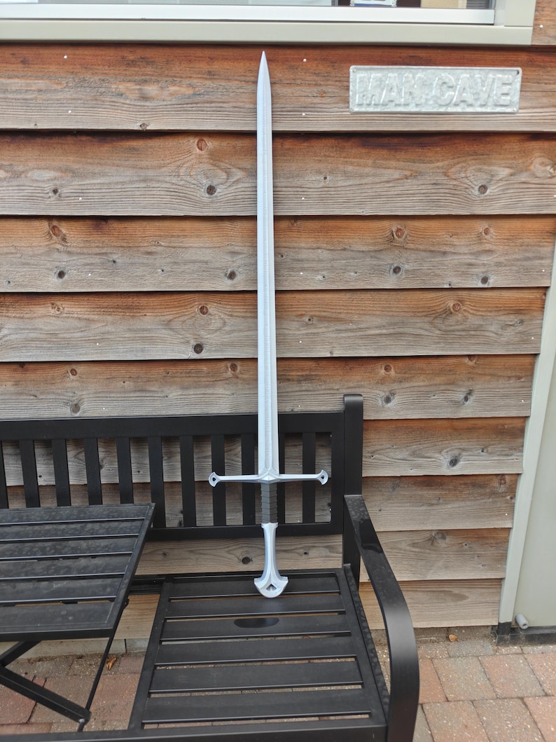 May include: A silver-colored sword with a long, pointed blade and a cross-shaped hilt. The sword is resting on a black metal bench in front of a wooden wall. The sign above the sword reads "MAN CAVE".