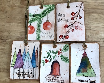 May include: Five handmade Christmas gift tags with watercolor illustrations. The tags feature a variety of holiday themes, including a Christmas tree, ornaments, holly, and a bell. The tags are decorated with the words "Winter Greetings", "Happy Holidays", "Season's Greetings", and "Greetings".