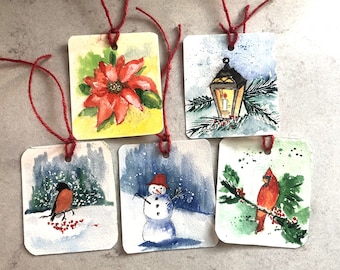 Könnte beinhalten: Satz von sechs handgemalten Geschenkanhängern in Aquarelltechnik mit winterlichen Weihnachtsmotiven. Die Anhänger zeigen eine rote Weihnachtsstern, einen Schneemann, einen Kardinal, einen Rotkehlchen, eine Laterne und eine Winterszene.