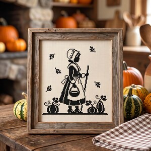 May include: A framed cross-stitch embroidery featuring a black silhouette of a person in a bonnet and apron holding a basket and staff, surrounded by falling leaves and pumpkins, set against a light beige fabric background in a rustic wood frame.
