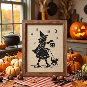 May include: A framed cross-stitch artwork on light beige fabric featuring a black silhouette of a witch wearing a pointed hat, holding a jack-o'-lantern, and walking a black cat under a crescent moon and stars, set in a simple wooden frame.