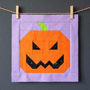 May include: A Halloween-themed fabric art piece featuring a geometric orange pumpkin face with black triangle eyes and a jagged mouth. The pumpkin has a green stem and is set against a lavender background. The artwork is hung with clothespins on a string.