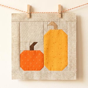 May include: A fabric art piece featuring two stylized pumpkins in shades of orange and yellow, with brown stems. The pumpkins are set against a neutral background and framed by a light beige border. The artwork is suspended by a string and wooden clothespins.