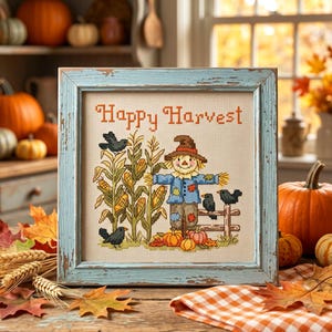Könnte beinhalten: Ein gerahmtes Kreuzstich-Stickbild mit dem Text Happy Harvest, einer Vogelscheuche, Maiskolben, Kürbissen und schwarzen Vögeln. Das Kunstwerk befindet sich in einem quadratischen, abgenutzten hellblauen Holzrahmen auf beigem Stoffgrund.