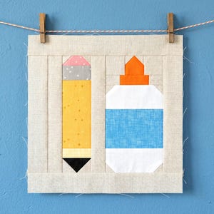 May include: A fabric art piece featuring a yellow pencil with a pink eraser and a white glue bottle with a blue label. The artwork is framed in a light beige fabric and is suspended by a clothesline and clothespins against a blue background.