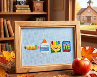 May include: A framed cross-stitch featuring school supplies: a pencil, glue bottle, crayon box, and calculator. The artwork is set against a light blue background, framed in light wood, and placed on a wooden table with fall leaves and an apple.