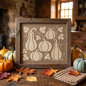 Peut inclure: Un cadre en bois carré contenant une broderie au point de croix sur tissu marron. La broderie présente un motif de cinq citrouilles et courges de couleur crème claire, entourées de vignes et de feuilles brodées sur un fond quadrillé.