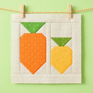 May include: A fabric quilt block featuring two carrot designs. The carrots are orange and yellow with green tops, set against a light beige background. The block is hung with wooden clothespins on a string against a light green backdrop.
