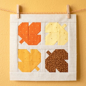 May include: A square fabric art piece featuring four stylized leaf designs in shades of orange, yellow, and brown. The leaves are set against a neutral background and framed with a light-coloured border. The artwork is hung with clothes pegs on a string.
