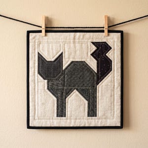 May include: A square, quilted wall hanging featuring a geometric black cat design. The cat is composed of various shades of gray fabric, with a black border and a cream-colored background. The artwork is suspended by clothespins on a black string.