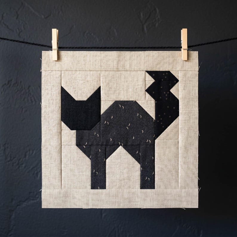 May include: A square fabric block featuring a geometric black cat design. The cat is pieced together with various black fabrics, some with small white accents. The block is framed in a neutral beige fabric and is suspended by a clothesline and wooden clothespins.