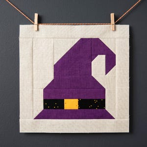 May include: A square fabric block featuring a purple witch's hat with a black band and yellow buckle. The hat is set against a cream-colored background and is suspended by a clothes pin and string, perfect for Halloween decor.