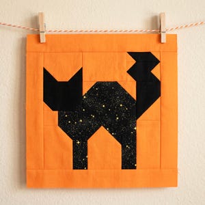 May include: A square, orange fabric block with a black cat design. The cat's body is filled with a starry pattern. The block is hung by a clothes pin on a string. The cat is made of geometric shapes.