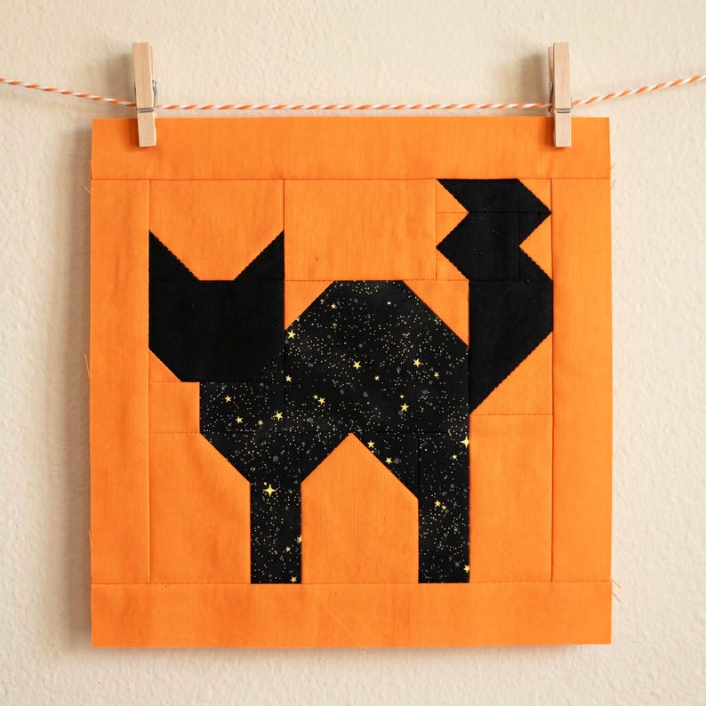 May include: A square, orange fabric block with a black cat design. The cat's body is filled with a starry pattern. The block is hung by a clothes pin on a string. The cat is made of geometric shapes.