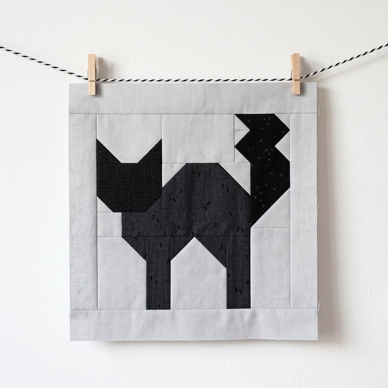 May include: A square quilt block featuring a geometric black cat design. The cat is constructed from various black fabrics with a light gray background and border. The quilt block is suspended by a clothes pin on a black and white string.