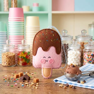 May include: A pink felt ice cream bar with a brown chocolate coating and colorful sprinkles. The ice cream bar has a smiling face and a wooden stick. The background includes jars of candy and ice cream toppings.
