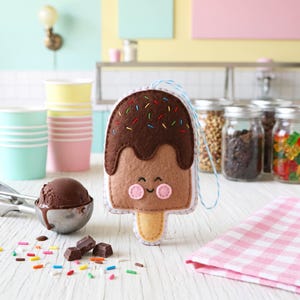 May include: A felt ice cream bar with a smiling face, brown chocolate topping, and colorful sprinkles. The ice cream bar has a light brown base and a wooden stick. A scoop of chocolate ice cream and chocolate pieces are nearby.