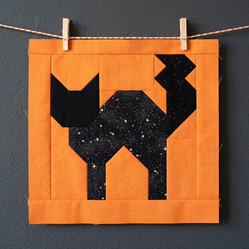 May include: A square, orange and black fabric block featuring a geometric black cat design. The cat's body is a starry black fabric, and the block is hung on a clothesline with clothespins. The design is likely for a quilt or other fabric craft.