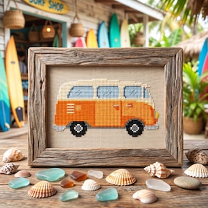 May include: Cross-stitch art of an orange and cream-colored vintage van in a rustic wooden frame. The artwork is displayed on a wooden table with seashells and sea glass. The background shows a surf shop and surfboards.