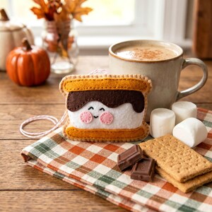 May include: A felt s'mores ornament with a smiling face, featuring a white marshmallow, dark chocolate, and graham cracker. The ornament is on a plaid napkin with a mug of coffee, marshmallows, and chocolate squares.