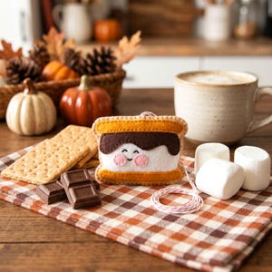 May include: A felt s'mores ornament with a smiling face, graham crackers, chocolate, and marshmallows on a plaid cloth. A mug of coffee, pumpkins, and pinecones are in the background. The ornament is approximately 3 inches wide.