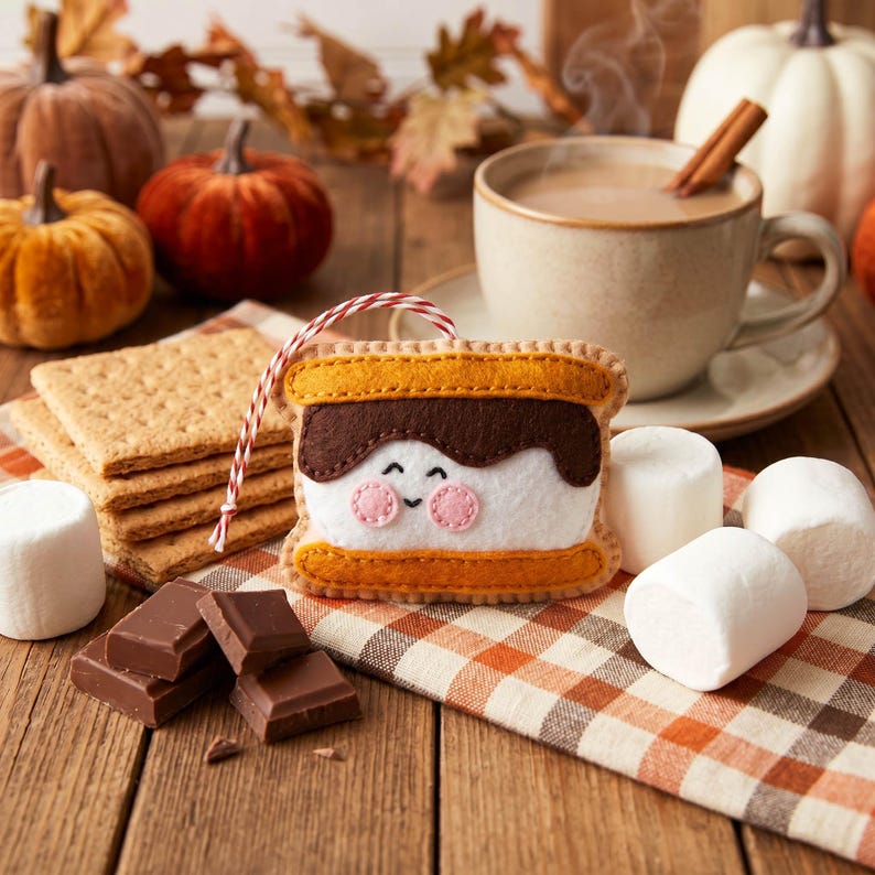May include: A felt s'mores ornament with a smiling face, graham crackers, chocolate pieces, and marshmallows. A cup of coffee with a cinnamon stick and pumpkins are in the background. The ornament is approximately 3 inches wide.