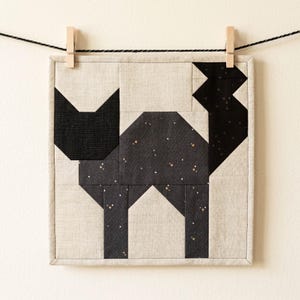 May include: A square, quilted wall hanging featuring a geometric black cat design. The cat is constructed from various fabric pieces, including a black fabric with gold stars. The background is a light beige fabric, and the piece is hung with clothespins.