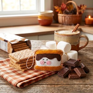 May include: A felt s'more ornament with a smiling face, graham crackers, marshmallows, and chocolate on a wooden surface. A mug of hot beverage with a cinnamon stick and a plaid napkin are also present. The scene evokes a cozy, autumnal theme.