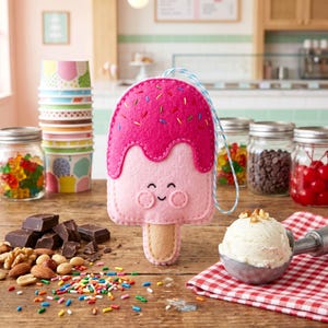 May include: A pink felt popsicle ornament with a smiling face and colorful sprinkles. The ornament is on a wooden surface with ice cream, toppings, and jars of candy. A fun, decorative item for an ice cream lover.