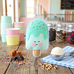 May include: A felt ice cream popsicle with a smiling face, mint green 'ice cream' and colorful sprinkles. The popsicle has a light brown stick and a blue and white string. The background includes ice cream cups, jars of toppings, and a scoop of ice cream.