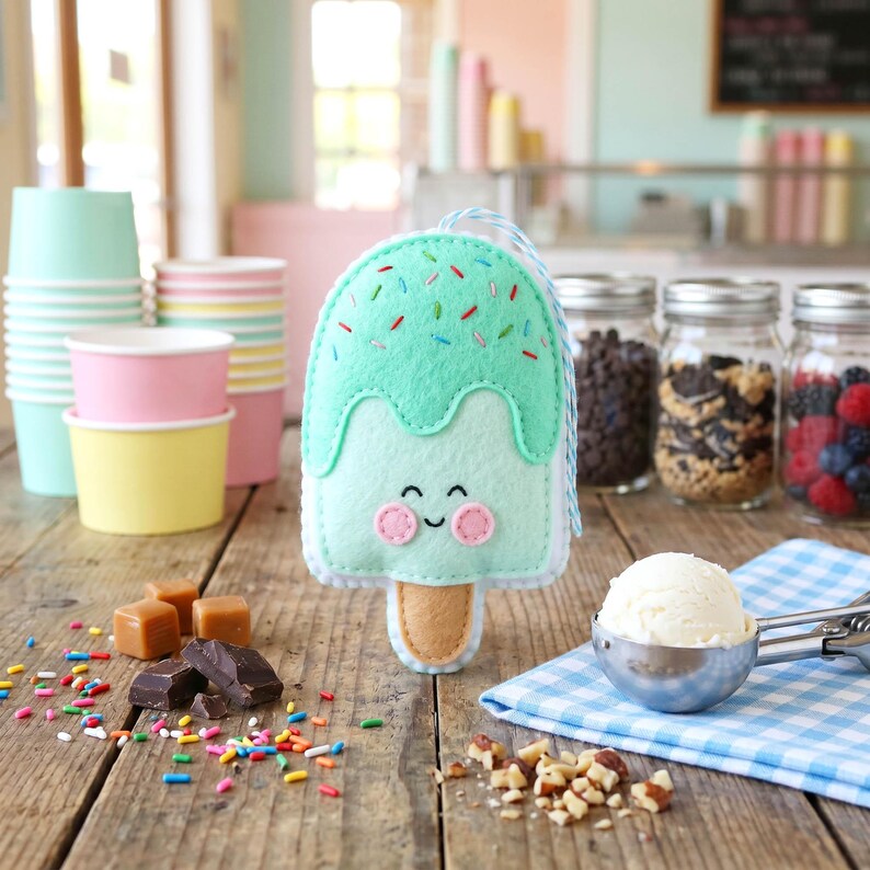 May include: A felt ice cream popsicle with a smiling face, mint green 'ice cream' and colorful sprinkles. The popsicle has a light brown stick and a blue and white string. The background includes ice cream cups, jars of toppings, and a scoop of ice cream.