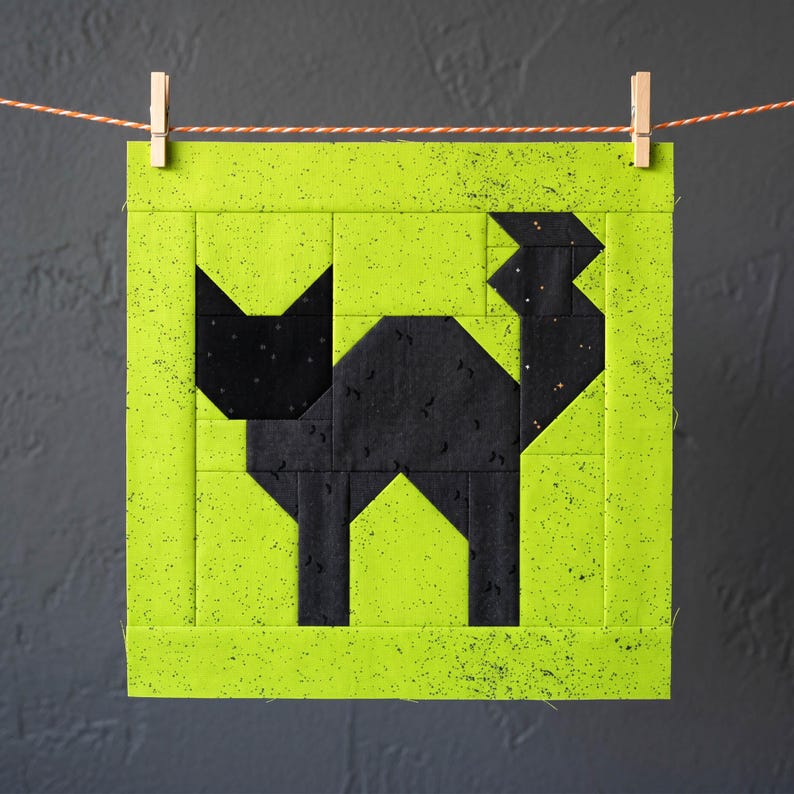 May include: A square fabric block featuring a black cat silhouette against a neon green background. The cat is constructed from geometric shapes. The block is suspended by a clothes pin on a string, suggesting a quilting or crafting project.