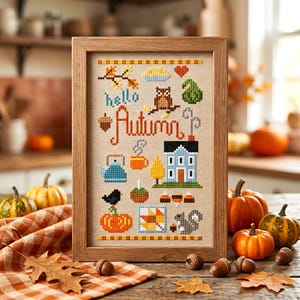 May include: A framed cross-stitch sampler on beige fabric featuring the text "hello Autumn" surrounded by seasonal motifs including an owl, a house, a pumpkin, a squirrel, a teapot, a mug, leaves, and candy corn in orange, blue, and brown.