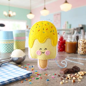 May include: A felt ice cream popsicle with a yellow top and a light yellow base. The popsicle has a smiling face with pink cheeks and is decorated with colorful sprinkles. A blue string is attached to the top.