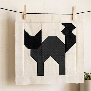 May include: A black and white geometric cat design on a square fabric panel. The cat is made of black fabric pieces, set against a cream-colored background. The panel is hung on a clothesline with wooden clothespins.