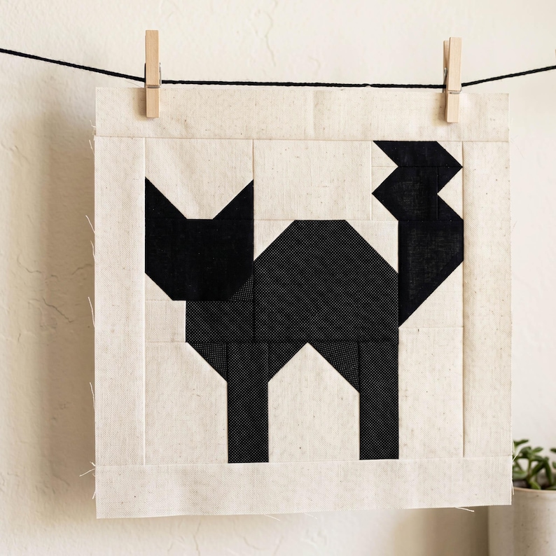 May include: A black and white geometric cat design on a square fabric panel. The cat is made of black fabric pieces, set against a cream-colored background. The panel is hung on a clothesline with wooden clothespins.