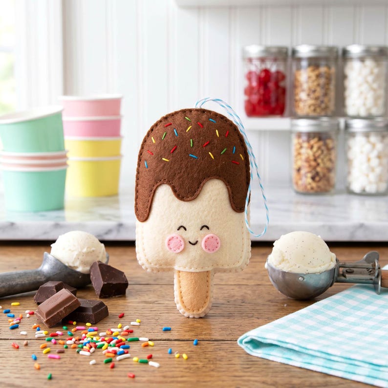 May include: A felt ice cream bar ornament with a smiling face, brown chocolate topping, and colorful sprinkles. The ornament has a wooden stick and a blue and white string for hanging. Ice cream scoops, chocolate pieces, and sprinkles are scattered on the wooden surface.