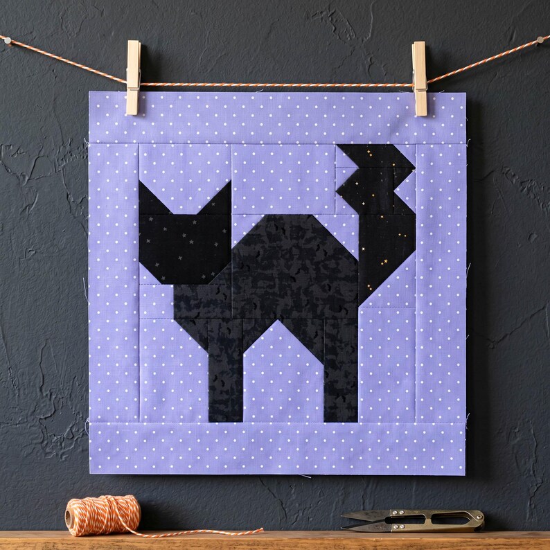 May include: A quilted square featuring a black cat design. The cat is made of various black fabrics and set against a lavender background with white polka dots. The quilt is hung with clothespins on a string.