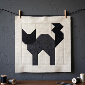 May include: A geometric quilt block featuring a black cat design. The cat is constructed from various black fabrics with a textured pattern, set against a neutral background. The artwork is displayed on a neutral fabric and hung with wooden clothespins.