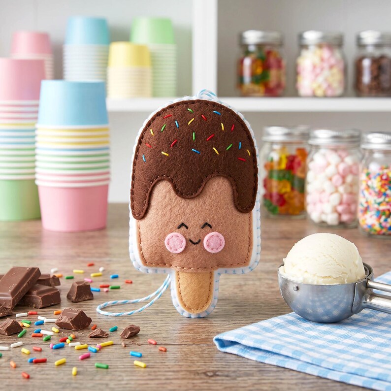 May include: A handmade felt ice cream bar with a smiling face, brown chocolate topping, and colorful sprinkles. The ice cream bar is on a wooden surface with chocolate pieces, colorful sprinkles, and a scoop of ice cream.