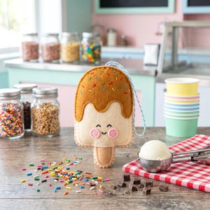 May include: A felt ice cream popsicle with a smiling face, brown 'chocolate' topping, and colorful sprinkles. The popsicle has a light blue string for hanging. Sprinkles and chocolate pieces are scattered on the wooden surface.