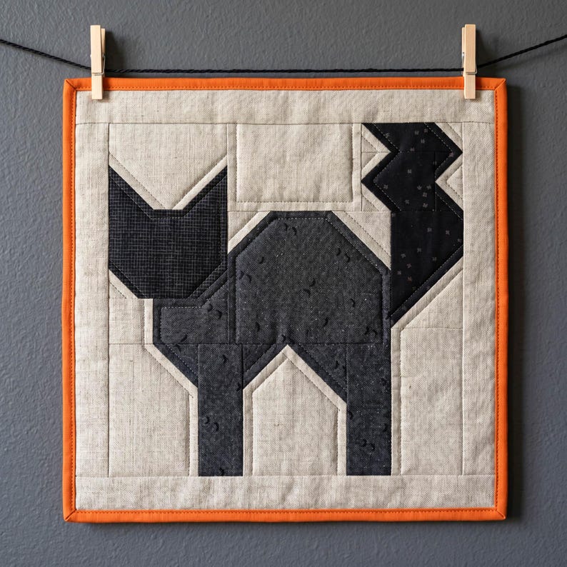 May include: A square, quilted wall hanging featuring a geometric black cat design. The cat is set against a neutral background with an orange border. The artwork is suspended by a clothesline and clothespins.