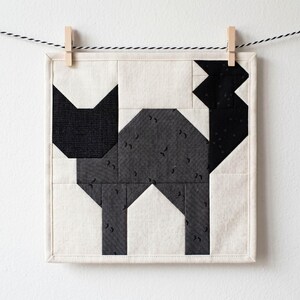May include: A square, quilted wall hanging featuring a geometric cat design. The cat is composed of black and gray fabric pieces on a cream background. The artwork is suspended by a clothes pin on a black and white string.